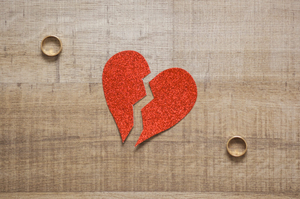 Broken red glitter heart on a wooden surface with two gold rings nearby.