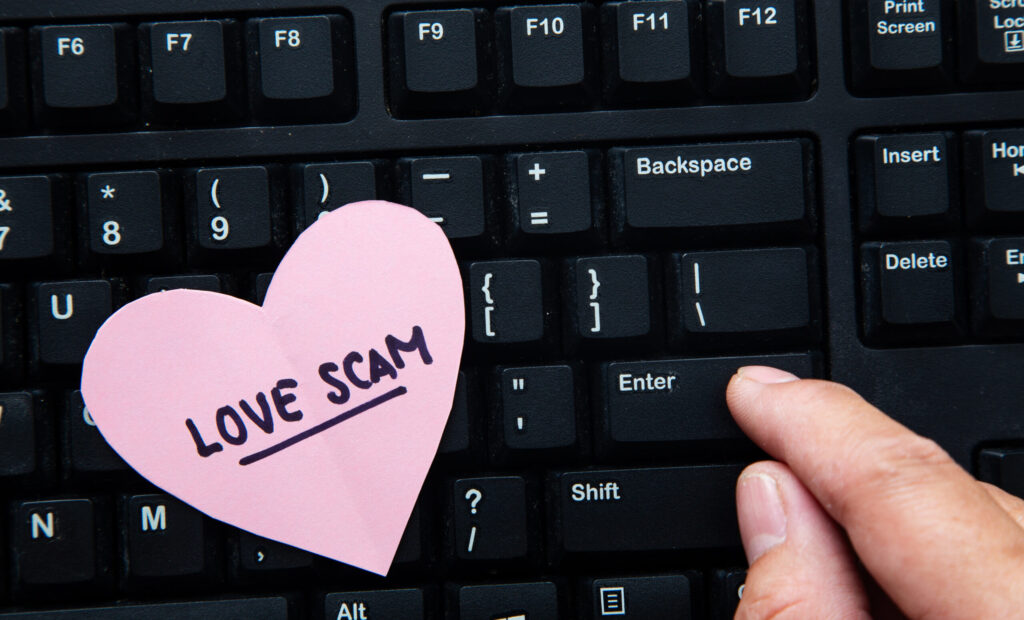 Close-up of a pink heart note reading 'LOVE SCAM' on a keyboard, with a finger near the Enter key.