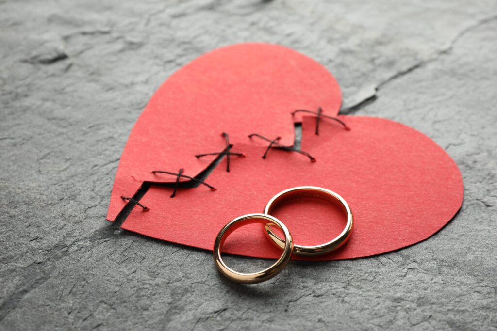 Red paper heart cutouts with black stitching and two gold wedding rings, set on a gray textured surface conveying love and commitment.