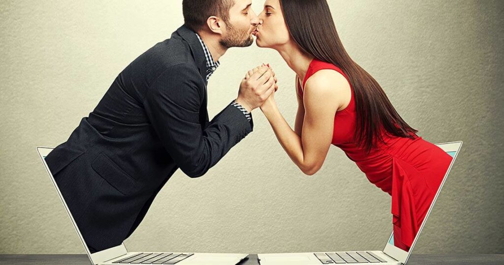 Couple kissing through two open laptops, hands clasped, symbolizing online romance or long-distance connection.