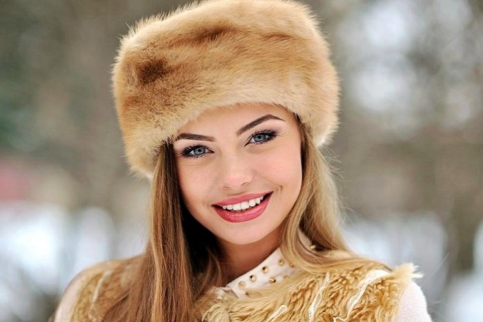 Young woman wearing a fluffy fur hat and beige coat, smiling outdoors in a snowy park.