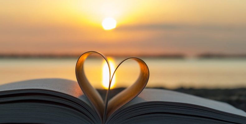 Open book on a beach at sunset, with the pages bent into a heart shape in the foreground.