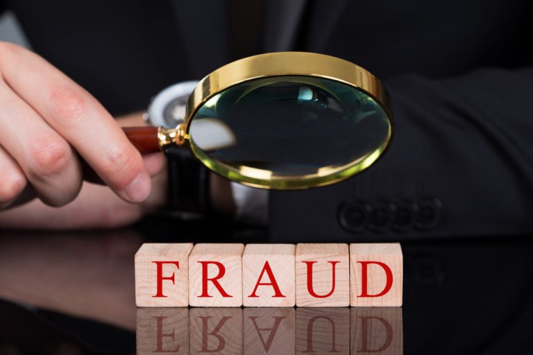 Person in a dark suit uses a magnifying glass to inspect wooden blocks spelling FRAUD.