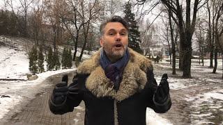 Man in a fur-collared coat speaking to the camera with hands raised in a snowy park setting.